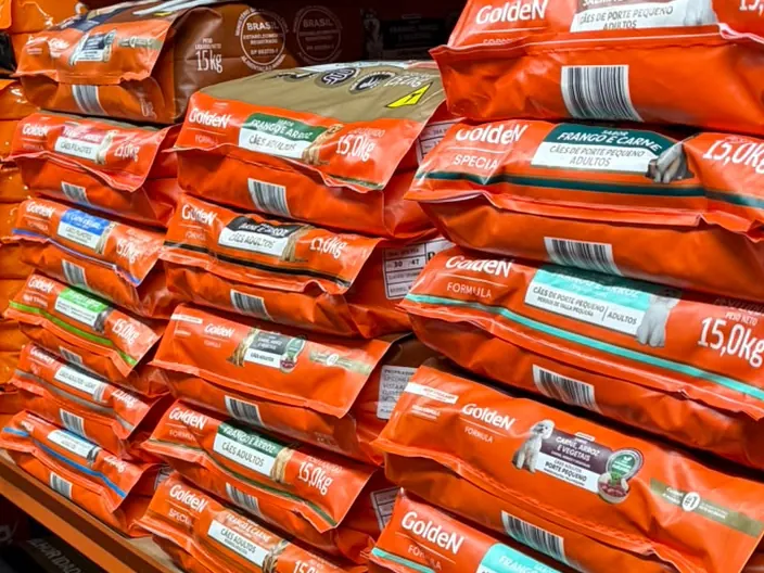 Prateleira da loja com sacos de ração empilhados; nas embalagens lê-se o peso 15,0 kg